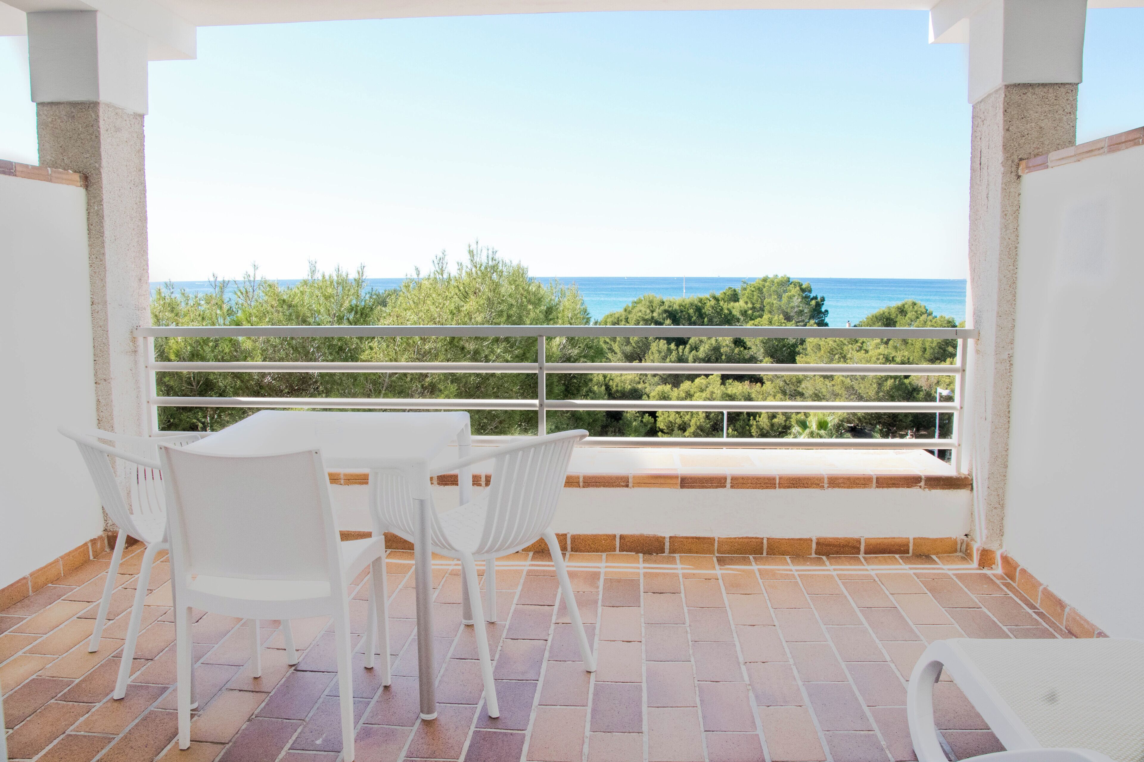 Un balcon avec des chaises blanches et une table, d’où l’on peut admirer une mer bleue et limpide ainsi qu’un paysage verdoyant.