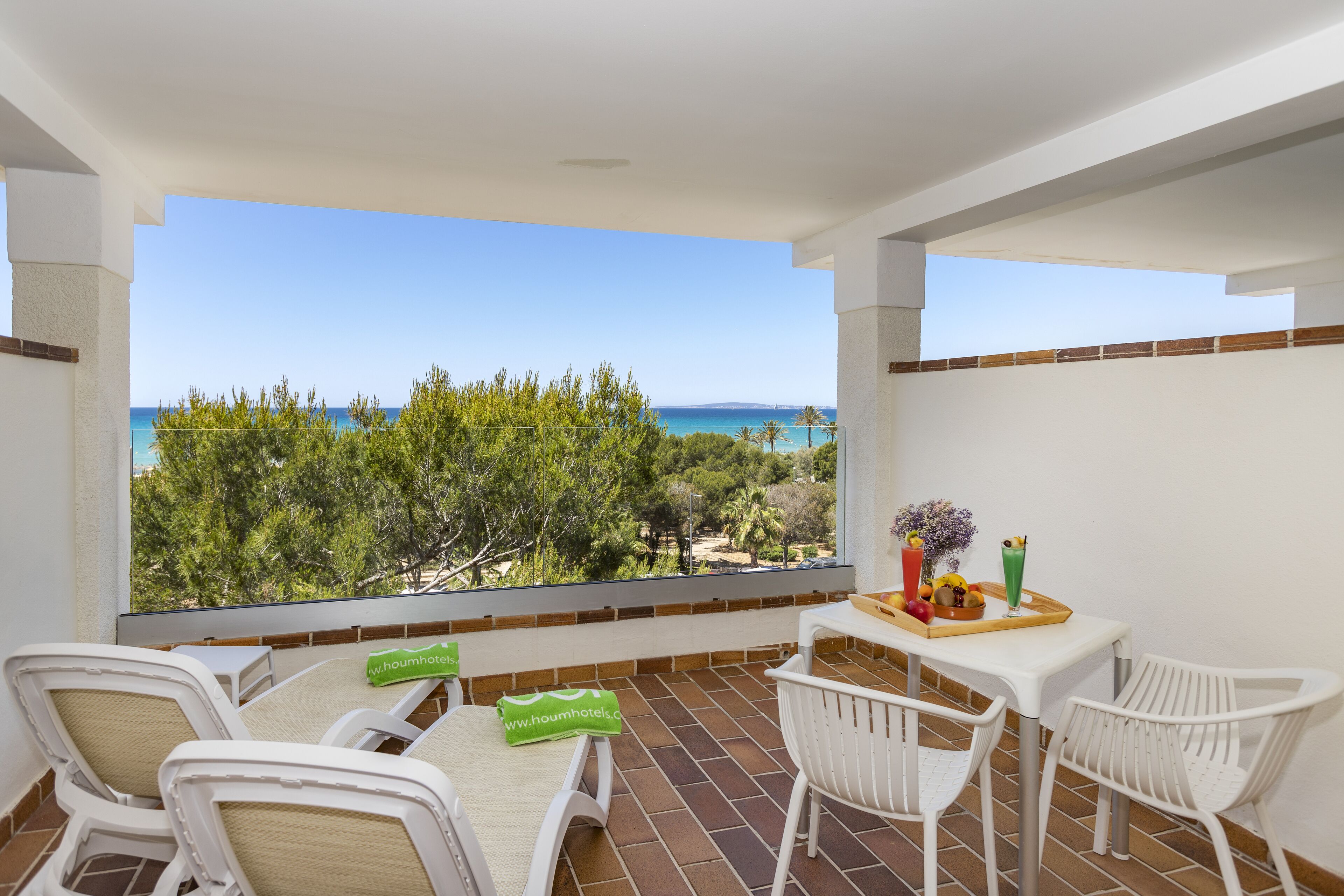 A balcony with a view of the sea, white chairs, a table with a tray of fruit, and a green towel.