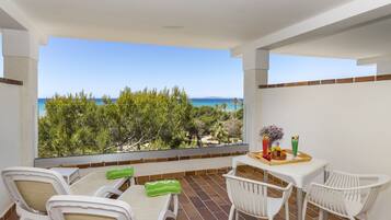 A balcony with a view of the sea, white chairs, a table with a tray of fruit, and a green towel.