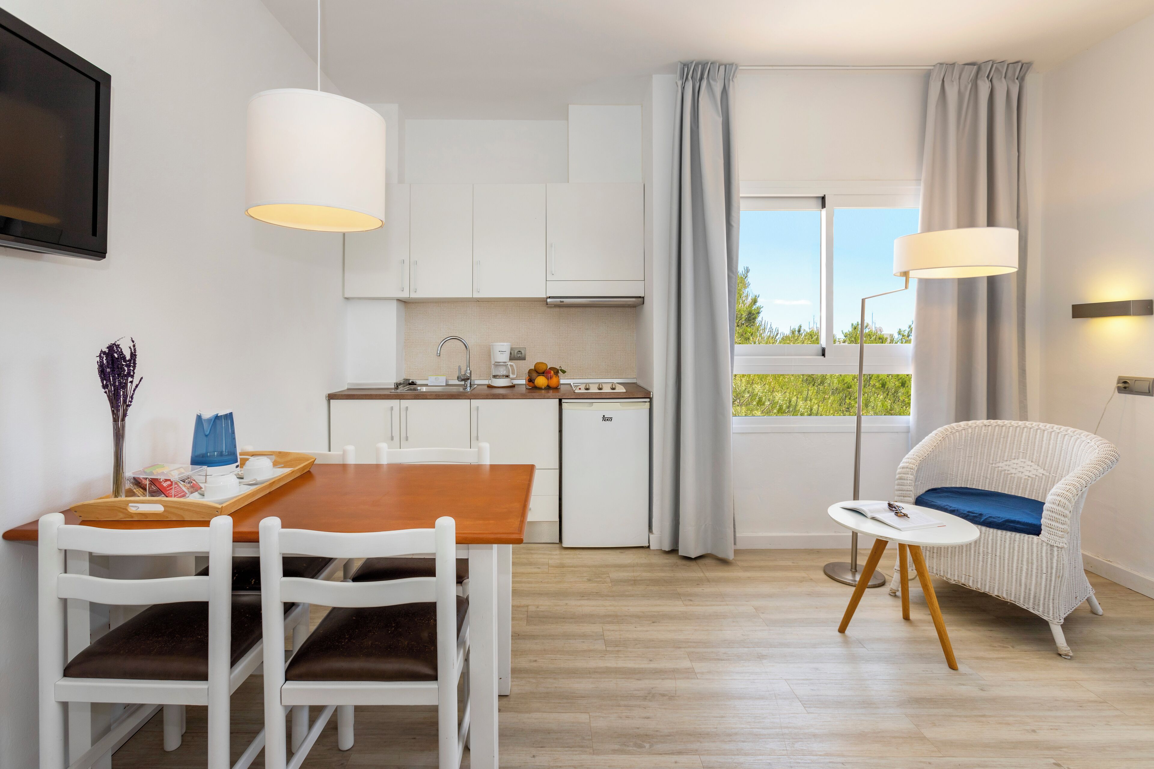 A modern kitchen with white cabinets, a dining table with chairs, a small refrigerator, and a window with curtains.
