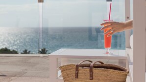 Une main tenant un verre avec une paille, un panier tressé, et une vue sur l’océan à travers une fenêtre en verre.
