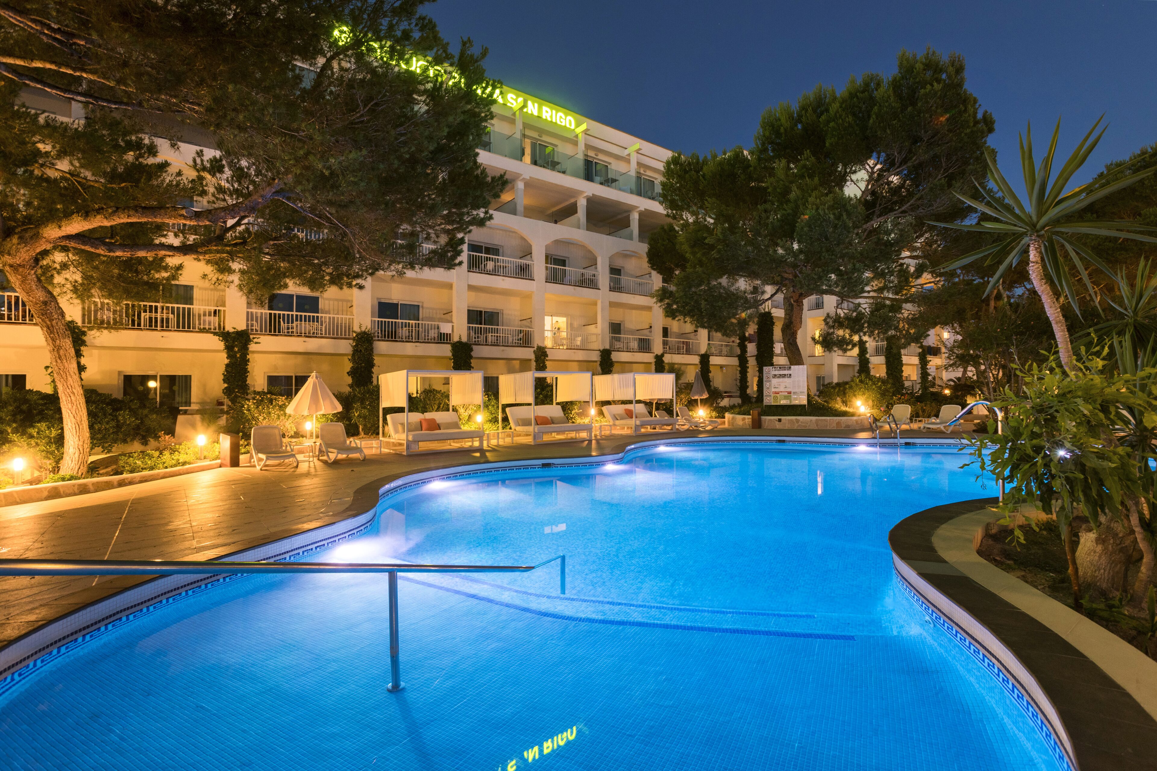 Una piscina con borde curvo, rodeada de sillones de descanso y sombrillas, con un edificio al fondo.