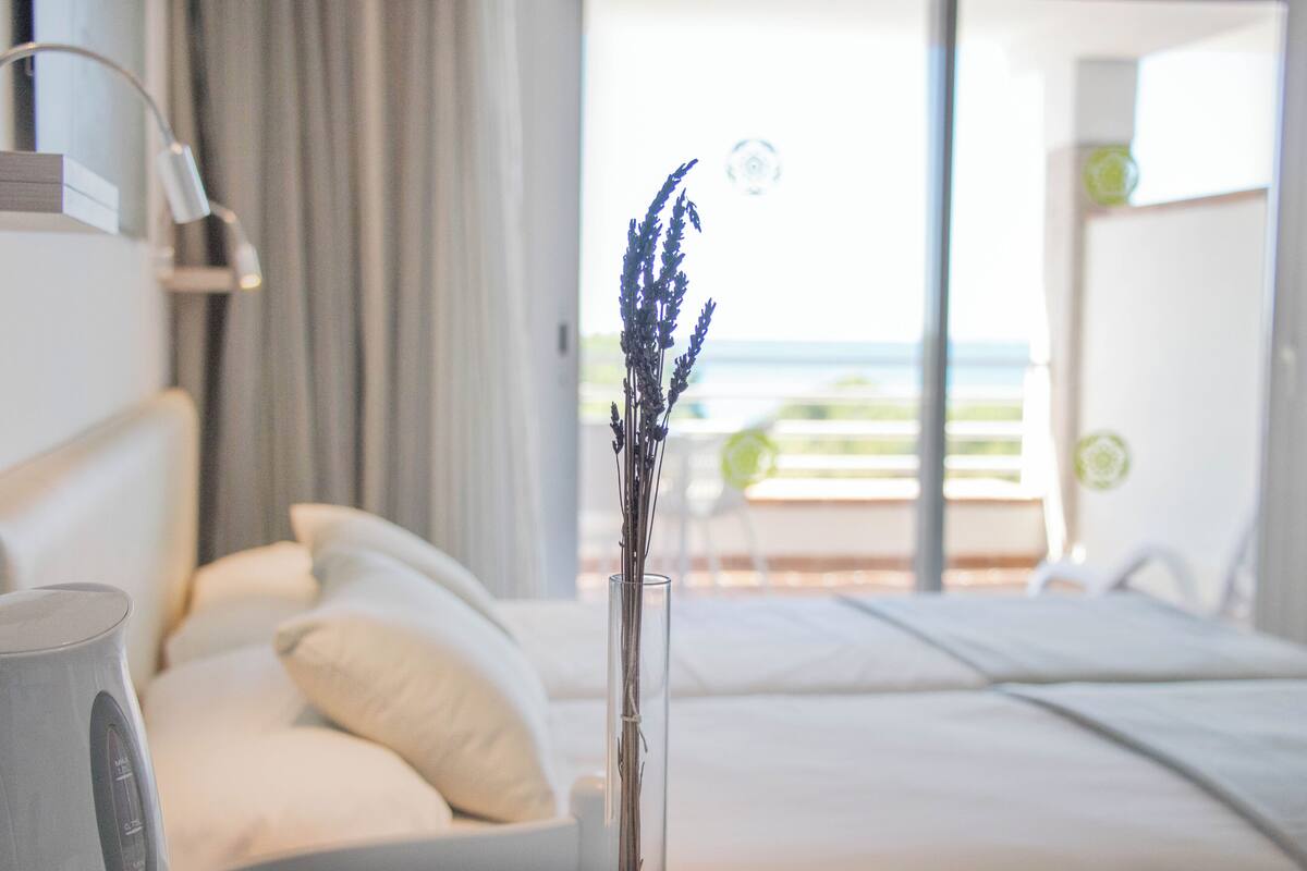 Habitación de hotel moderna con cama, mesita de noche con lámpara y vista al mar a través de un ventanal grande.