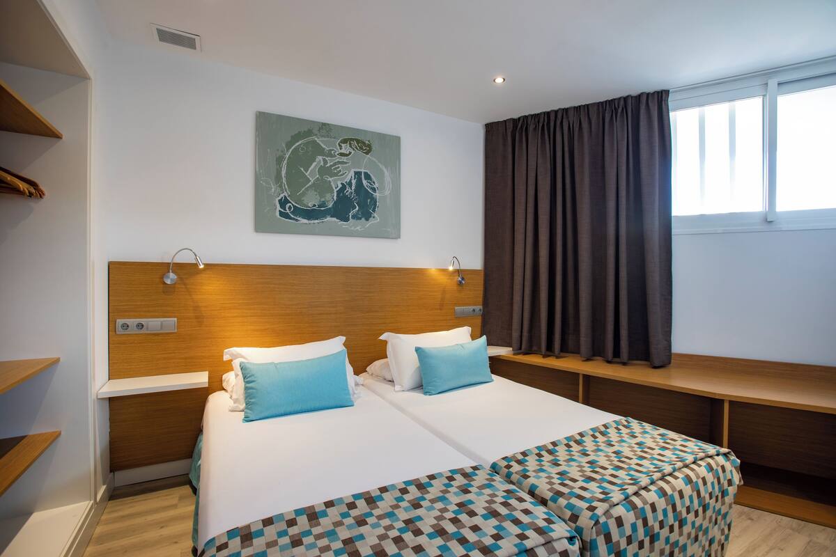 Habitación de hotel con dos camas, cabecera de madera, una obra de arte colgada en la pared, ventana con cortinas y una mesita de noche con lámpara.