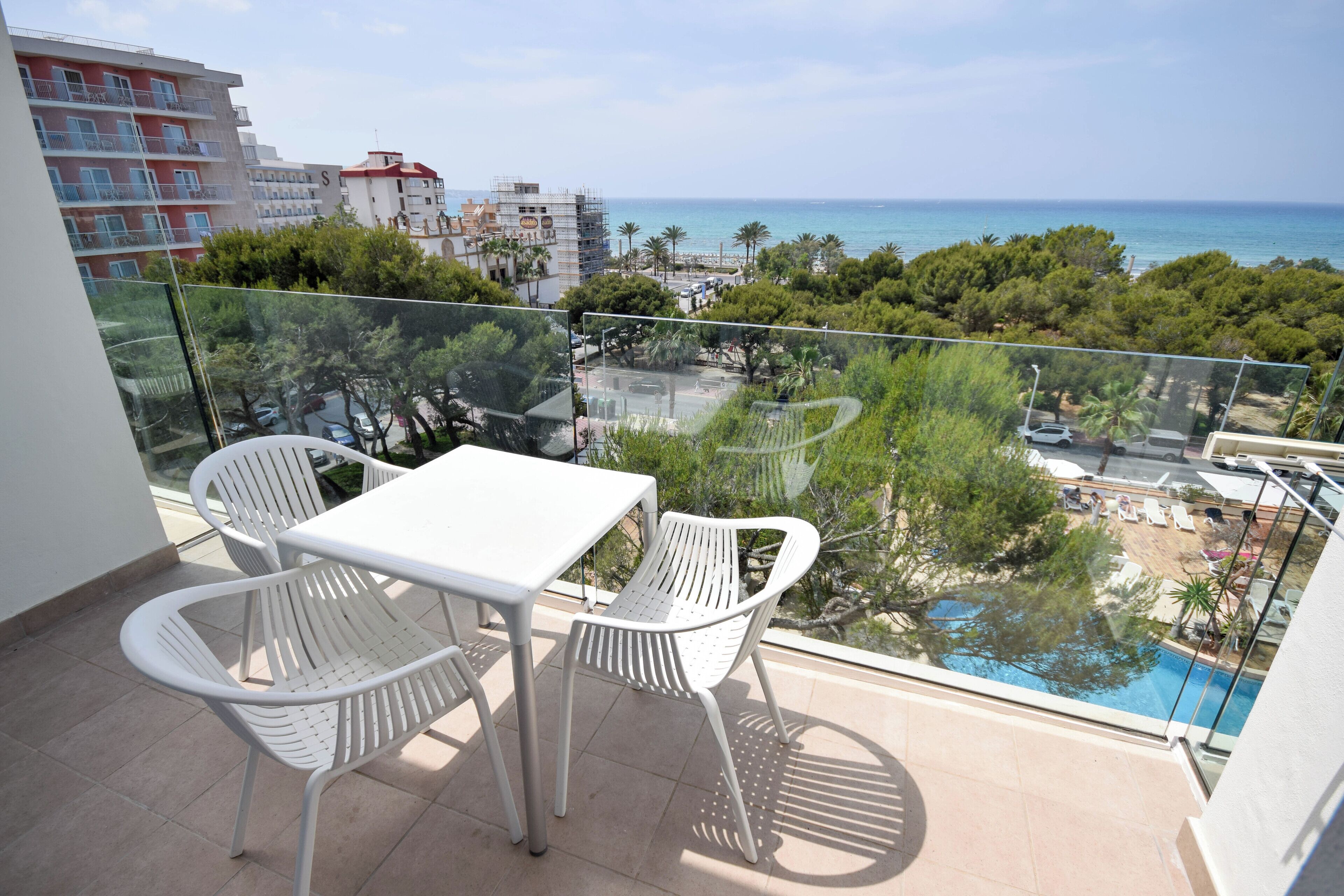 Un balcón con mesa y sillas, con vista a una piscina y una zona de playa con palmeras y edificios al fondo.