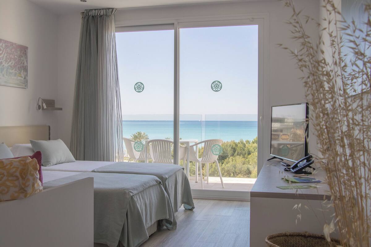 Een hotelkamer met twee bedden, een balkon met uitzicht op zee, en een bureau met een computer en papieren.