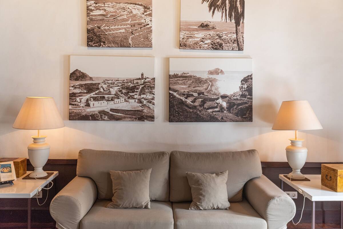 A living room with a beige sofa, two side tables with lamps, and four black-and-white photographs on the wall.