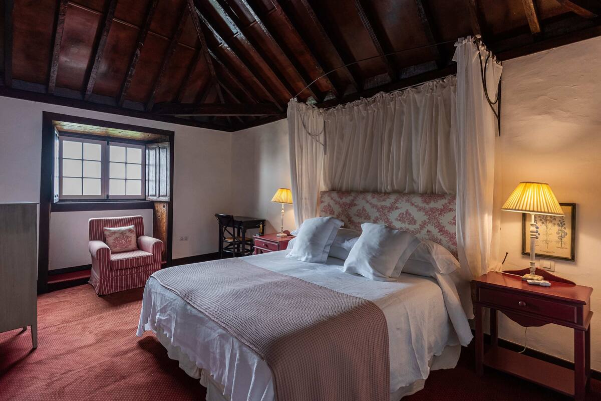 A bedroom with a large bed, a chair, a desk, and a window.