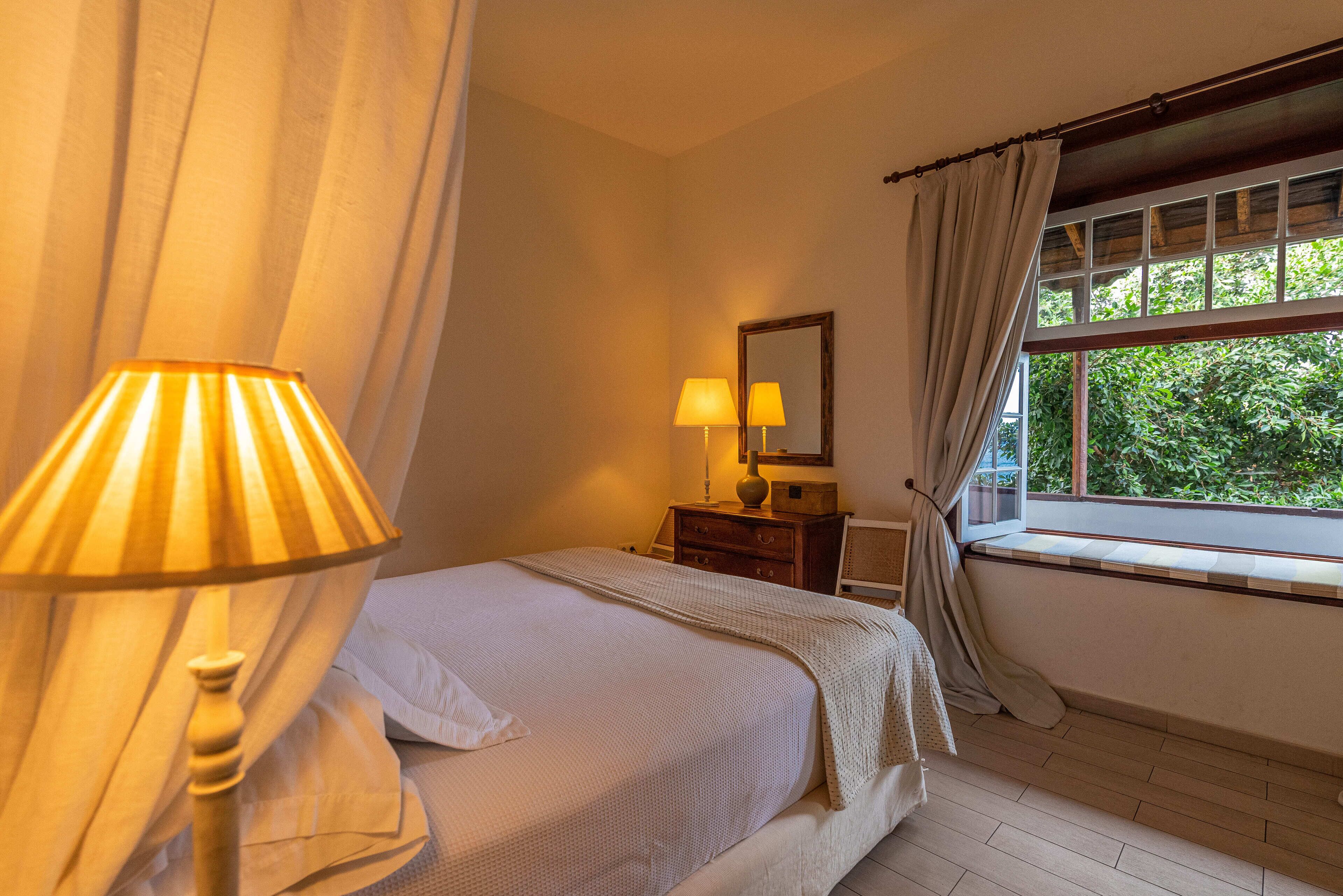 A bedroom with a bed, bedside lamps, a nightstand, a window with curtains, and a view of greenery outside.