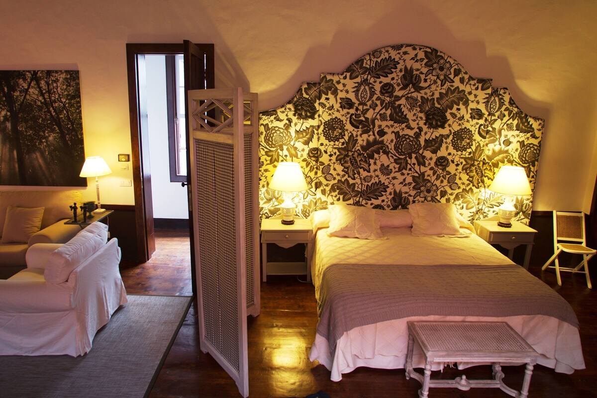 A bedroom with a large bed, two bedside tables with lamps, a chair, and a painting on the wall.