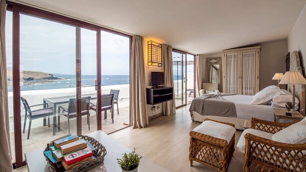 A hotel room with a large sliding glass door to the beach, a bed, a TV, wicker furniture, and a table with books and a plant. - Isla Baja Suites (Garachico)