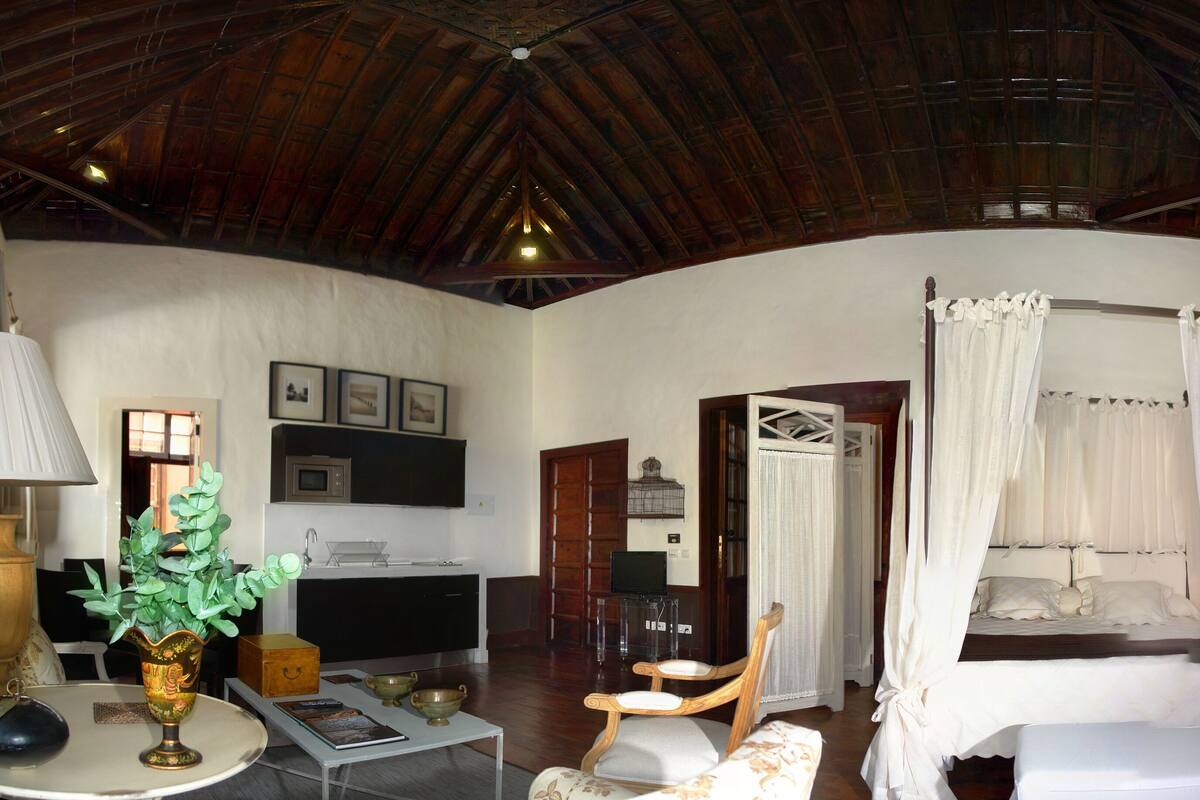 A room with a wooden ceiling, a bed with white curtains, a wooden chair, a table with a vase, and shelves with baskets.