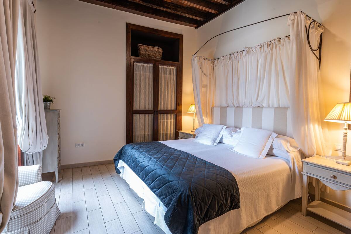 A bedroom with a large bed, a bedside table with a lamp, a chair, and a window with curtains.