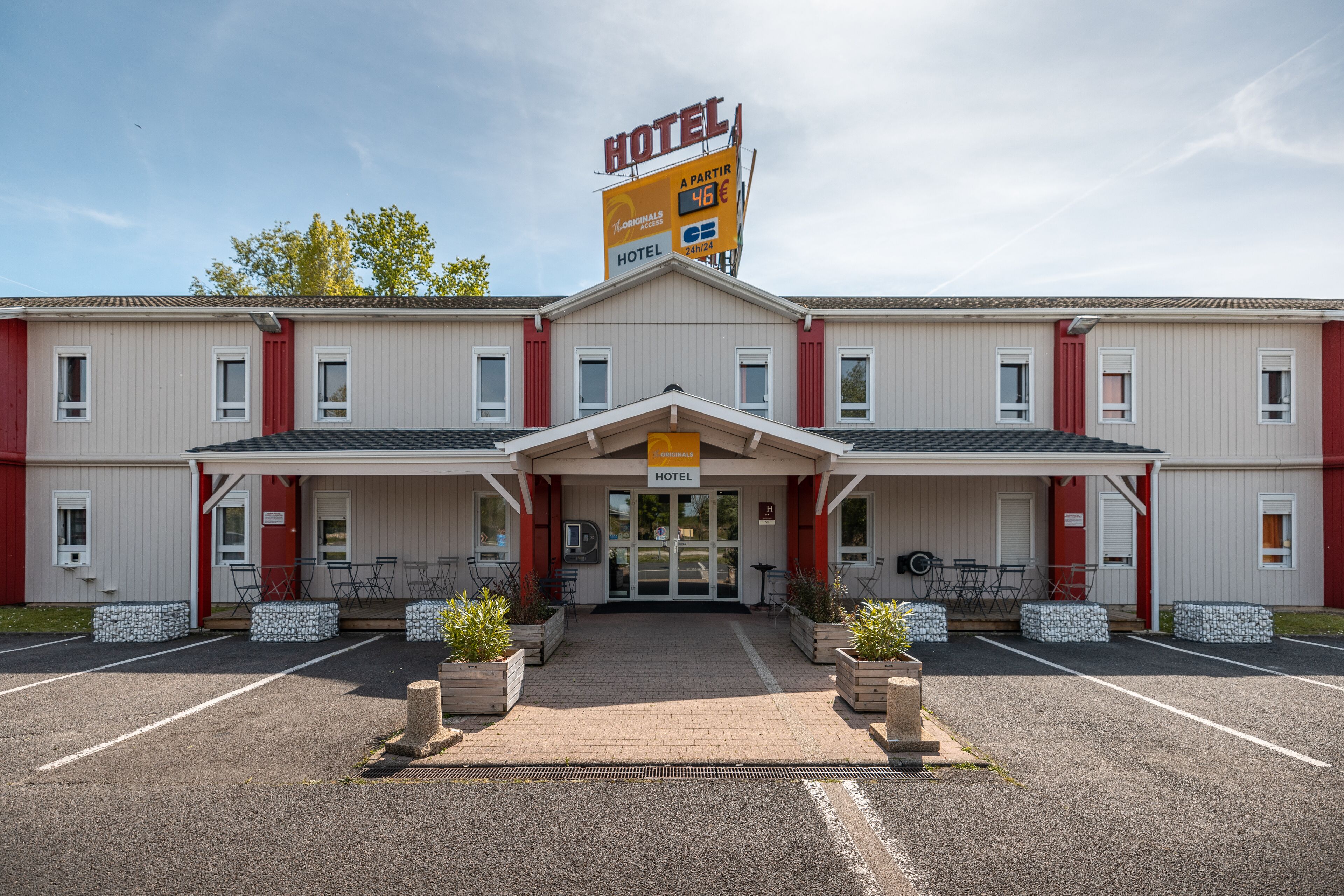 Un hôtel à deux étages avec une entrée centrale, des places assises à l’extérieur et un panneau indiquant un prix de 46 €.