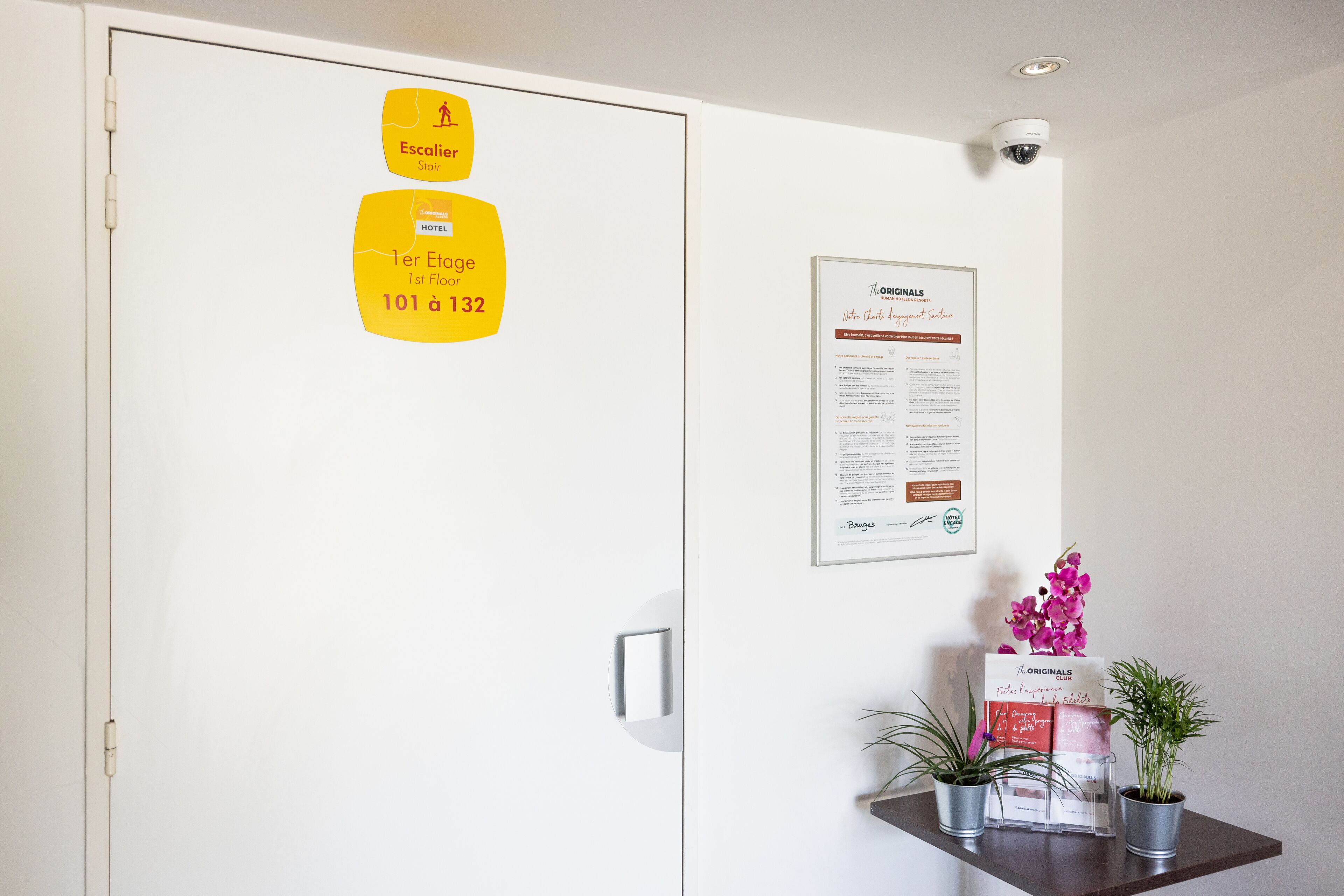 A hotel room with a door marked 'Escalier' and '1st Floor 101 à 132'. There is a framed document on the wall and two potted plants on a shelf.