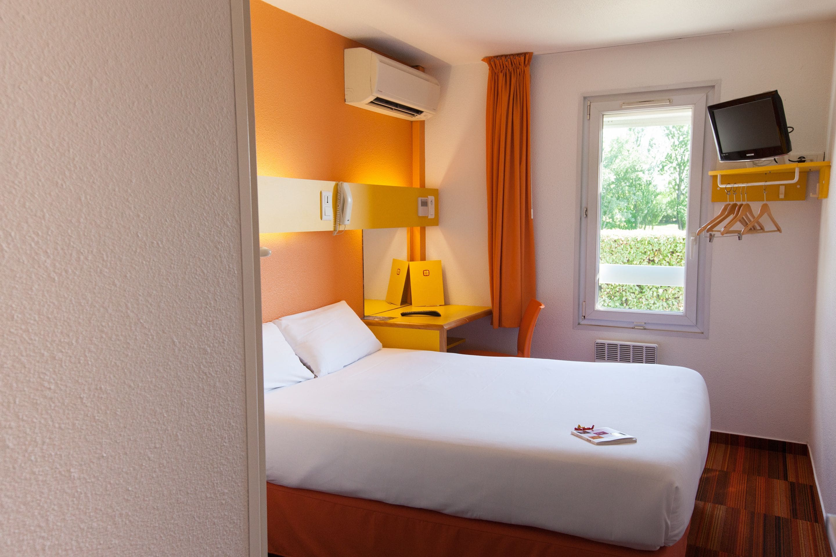 Quarto de hotel com cama, escrivaninha com cadeira, ar condicionado, televisão numa prateleira e janela com cortinas.