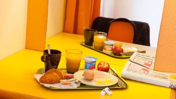 Ett frukostbricka med croissant, juice, yoghurt, ägg, skinka och ett äpple på ett gult bord.
