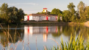 A building with a red and white facade, situated by a calm lake with reflections. - The Originals Access, Hôtel Bordeaux Lac (Bruges)