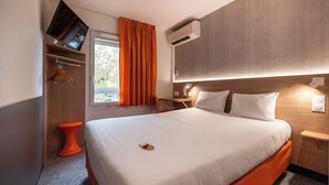A hotel room with a bed, a TV mounted on the wall, an air conditioning unit, a small orange stool, and a window with orange curtains. - The Originals Access, Hôtel Bordeaux Lac (Bruges)