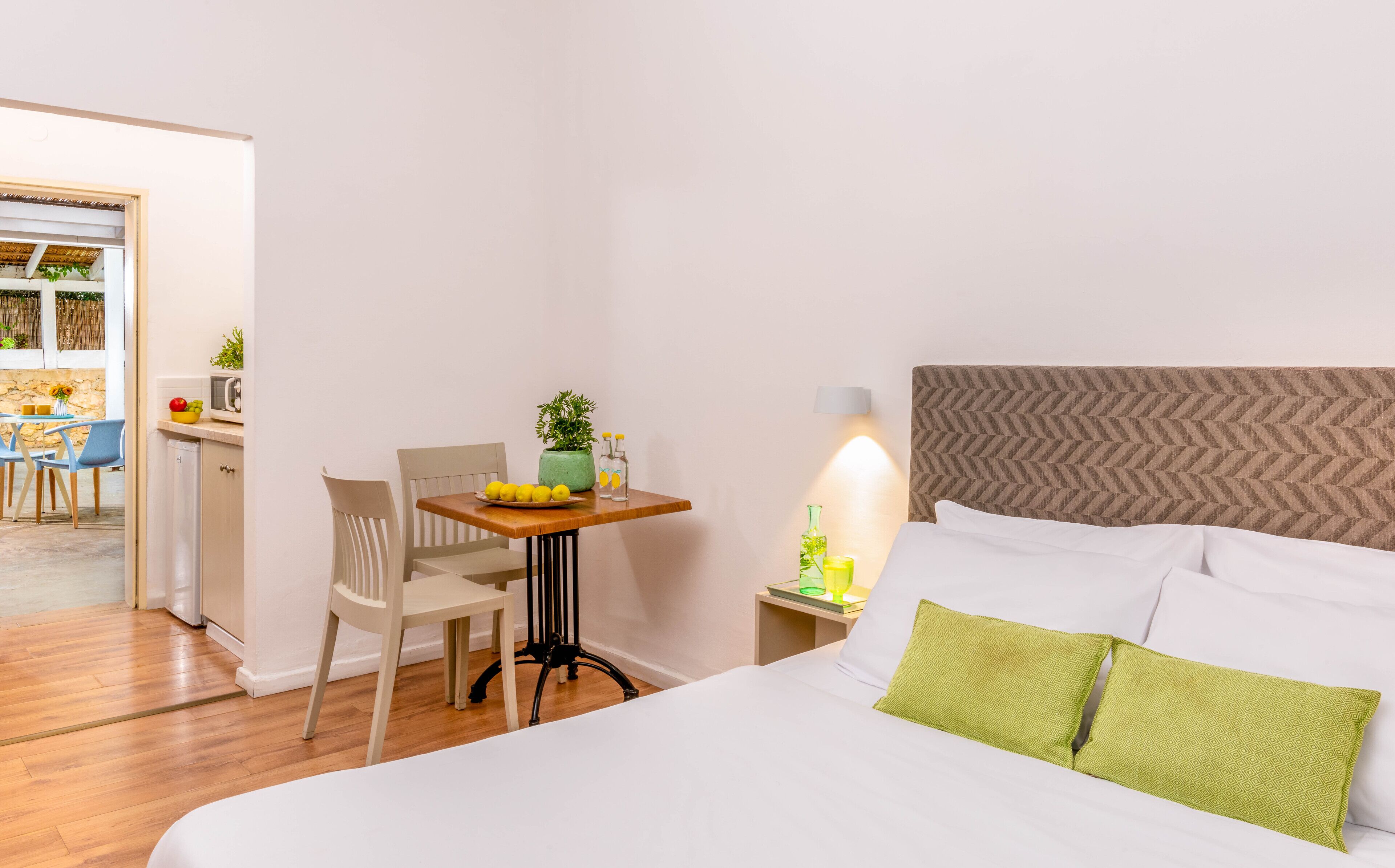 A hotel room with a bed, a bedside table with a lamp, a chair, and a small table with a bowl of fruit and a plant.