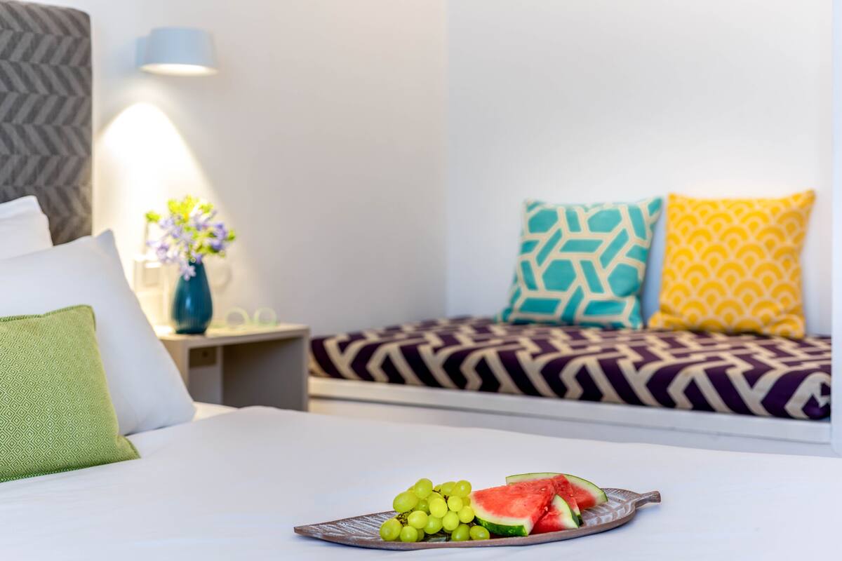 A hotel room with a bed, a bedside table with a vase of flowers, and a tray with grapes and watermelon.