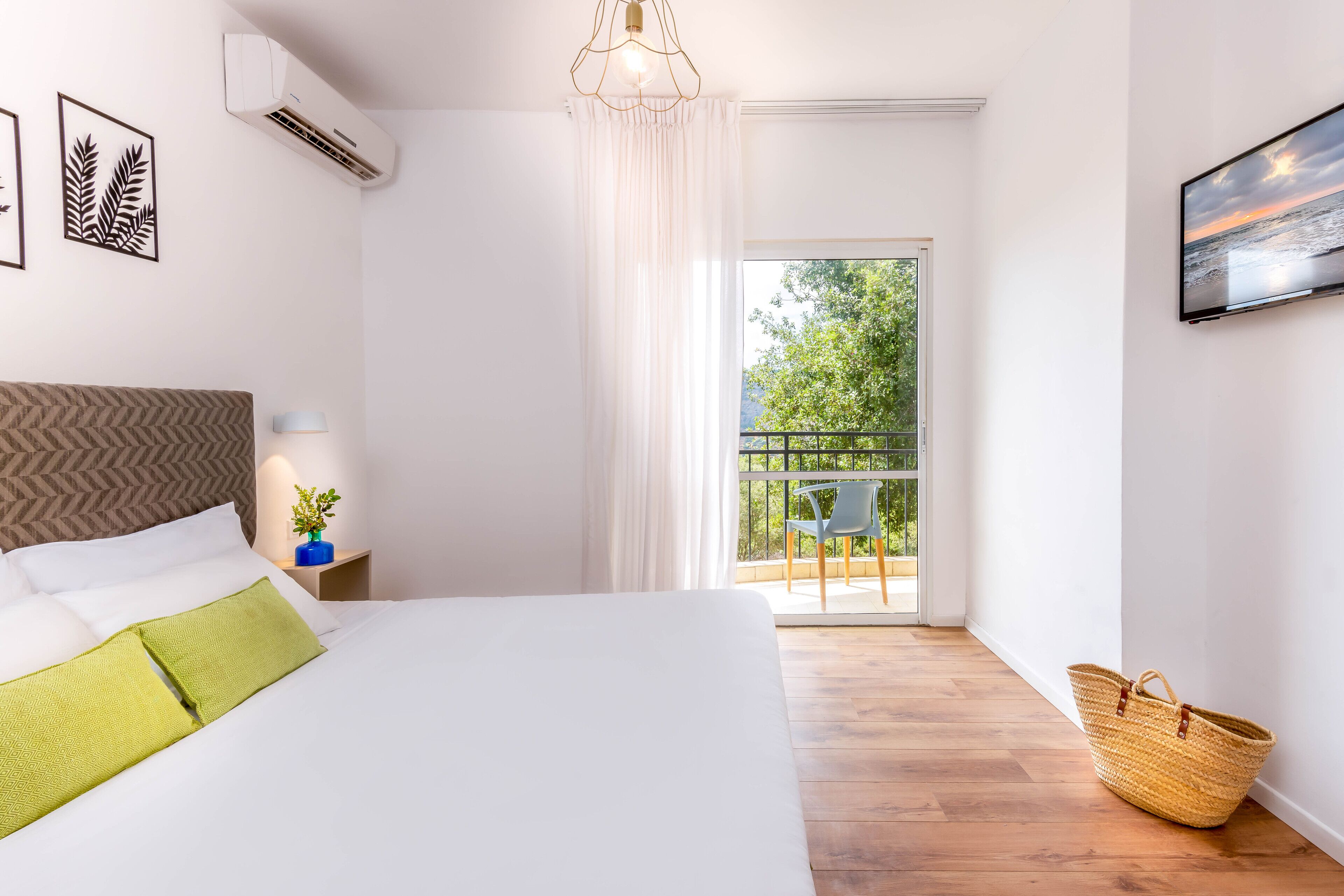 Un dormitorio con cama, mesita de noche, lámpara, balcón con silla y televisor montado en la pared.