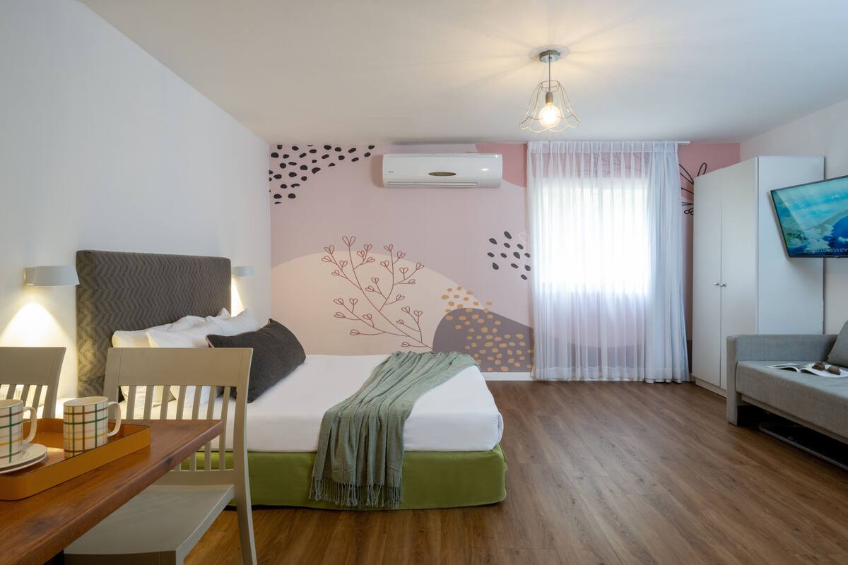 A modern bedroom with a bed, a desk with a tray of mugs, a chair, a TV, and an air conditioning unit.