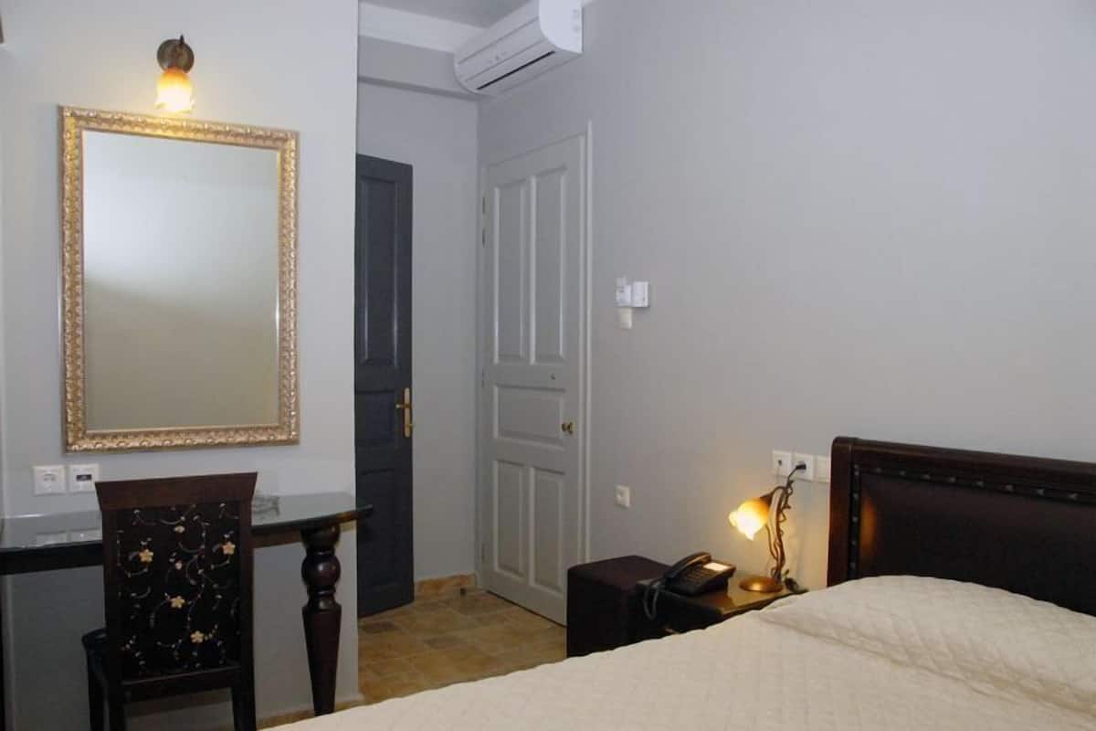 A hotel room with a bed, a desk with a chair, a mirror, a bedside table with a lamp, and a door.