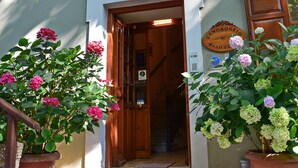 A building entrance with a wooden door, a black mat, and potted plants with pink flowers. - Filoxenia (Volos)