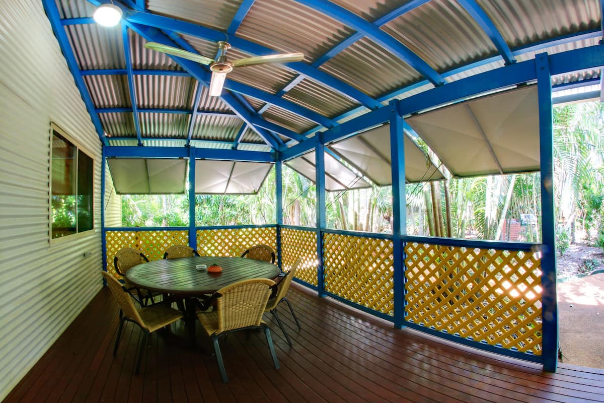 A covered outdoor area with a table and chairs, a ceiling fan, and a view of greenery outside.