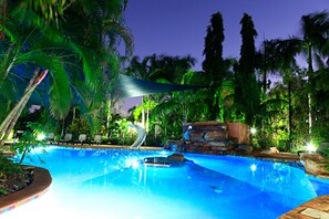 A swimming pool with lounge chairs, a water slide, and palm trees at dusk.