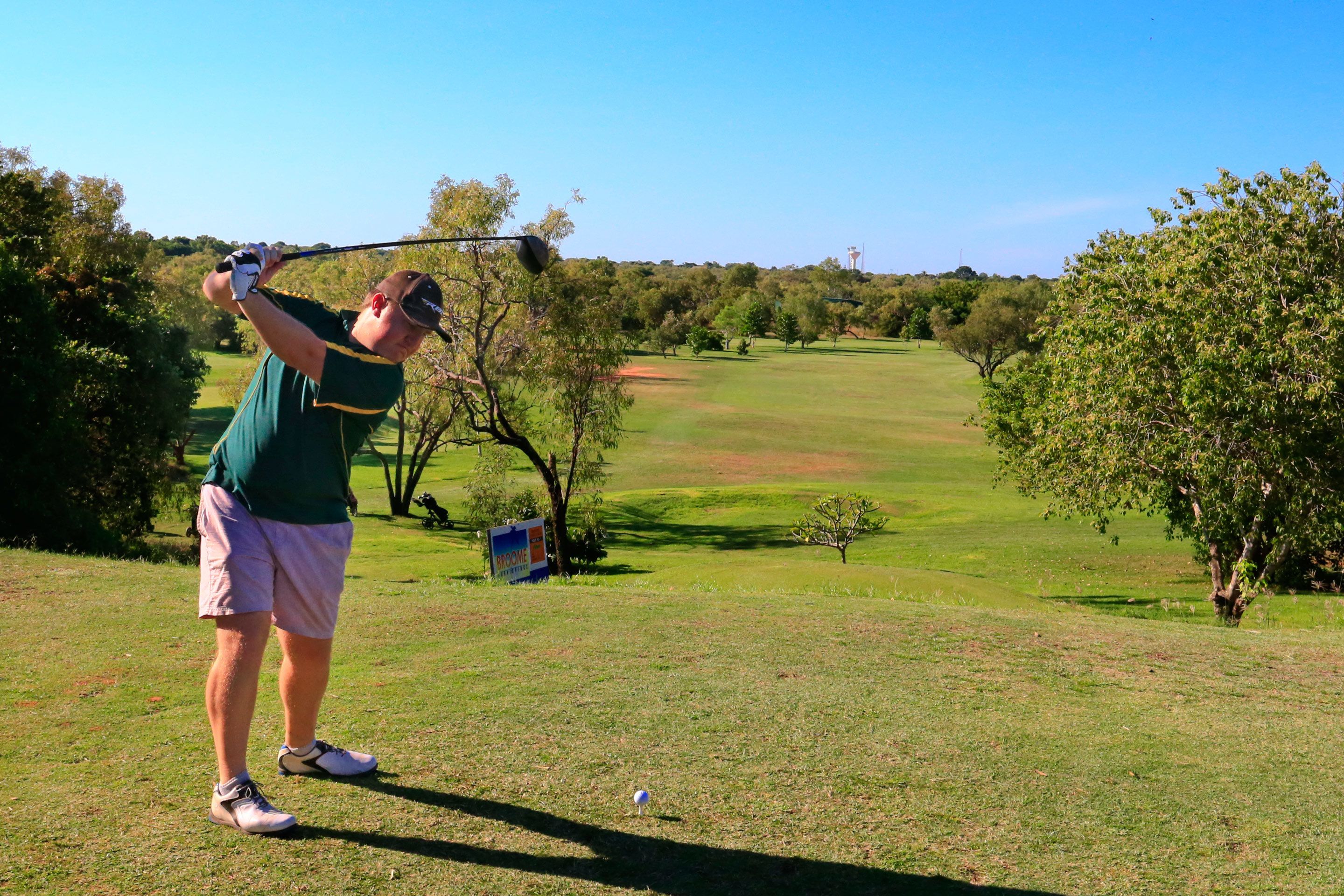 A golf course with a player in mid-swing, trees, and a clear sky.