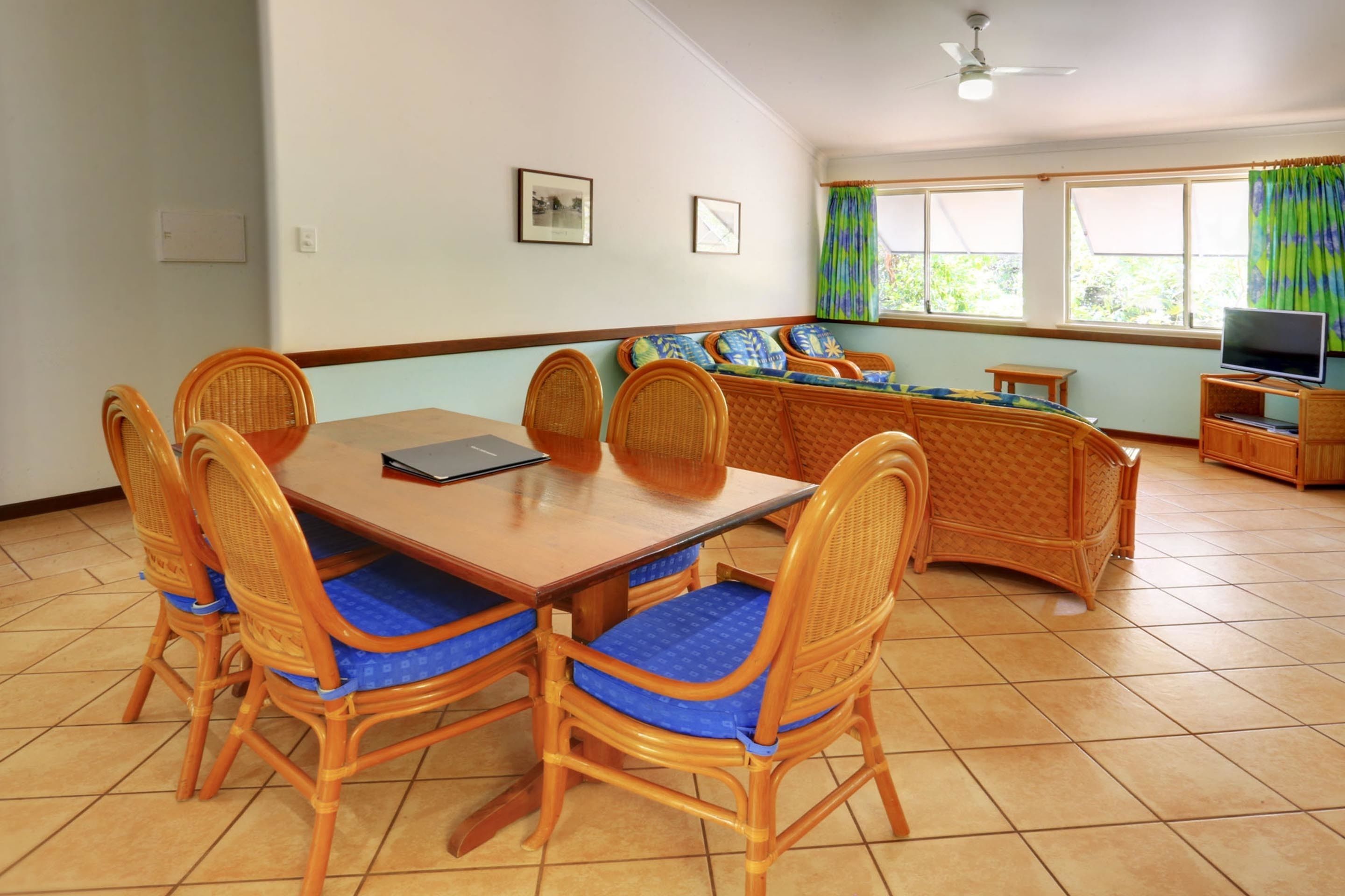 A dining area with a wooden table and wicker chairs, a sofa with cushions, and a television on a wooden stand.