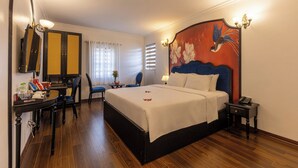 A hotel room with a large bed, a desk with a chair, a small table with a lamp, and a painting on the wall. - Hanoi House Hotel & Travel (Hanoi)