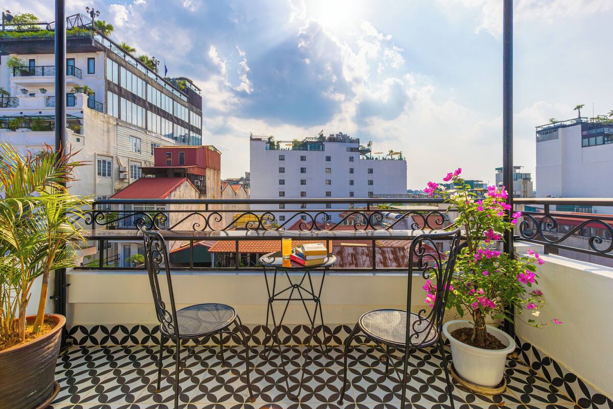 Un balcon doté d’un sol à motifs, de deux chaises, d’une table avec un livre et un verre, et d’un pot de fleurs contenant des fleurs roses.