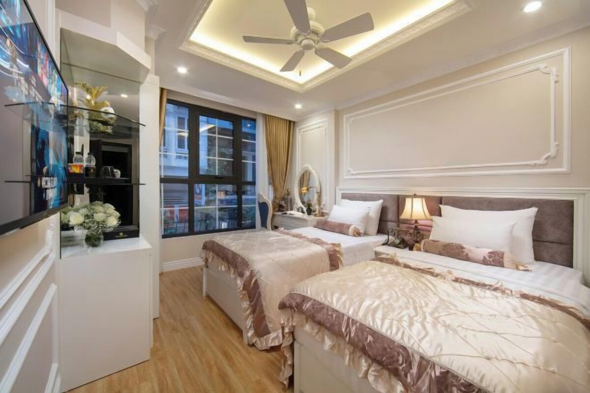 Camera d'albergo con due letti, una ventola a soffitto, un'ampia finestra con tende, un tavolino con lampada e una vetrina in vetro con fiori.