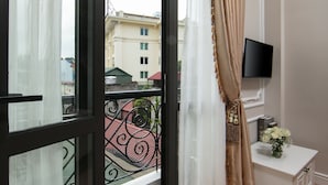 Un balcon donnant sur des immeubles, un téléviseur fixé au mur et une petite table sur laquelle est posé un vase de fleurs.