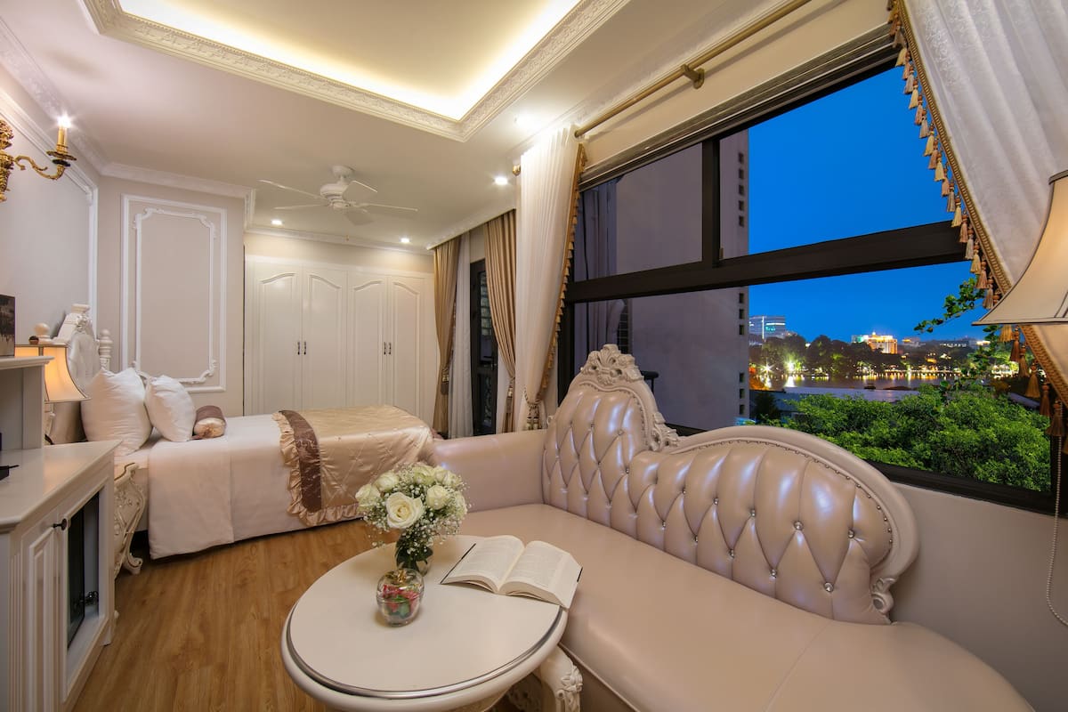 A well-lit bedroom with a large window, a sofa, a bedside table, and a bed with a headboard.