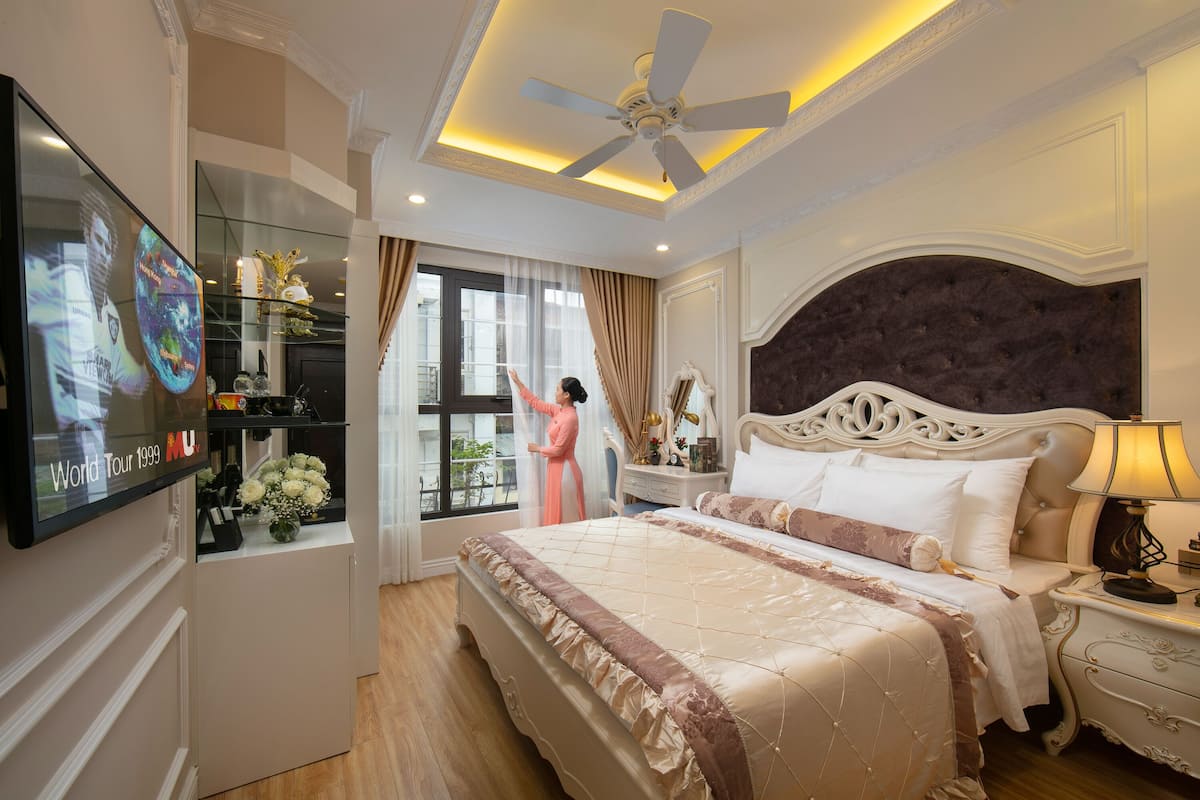 A bedroom with a large bed, a ceiling fan, a TV mounted on the wall, and a window with curtains.