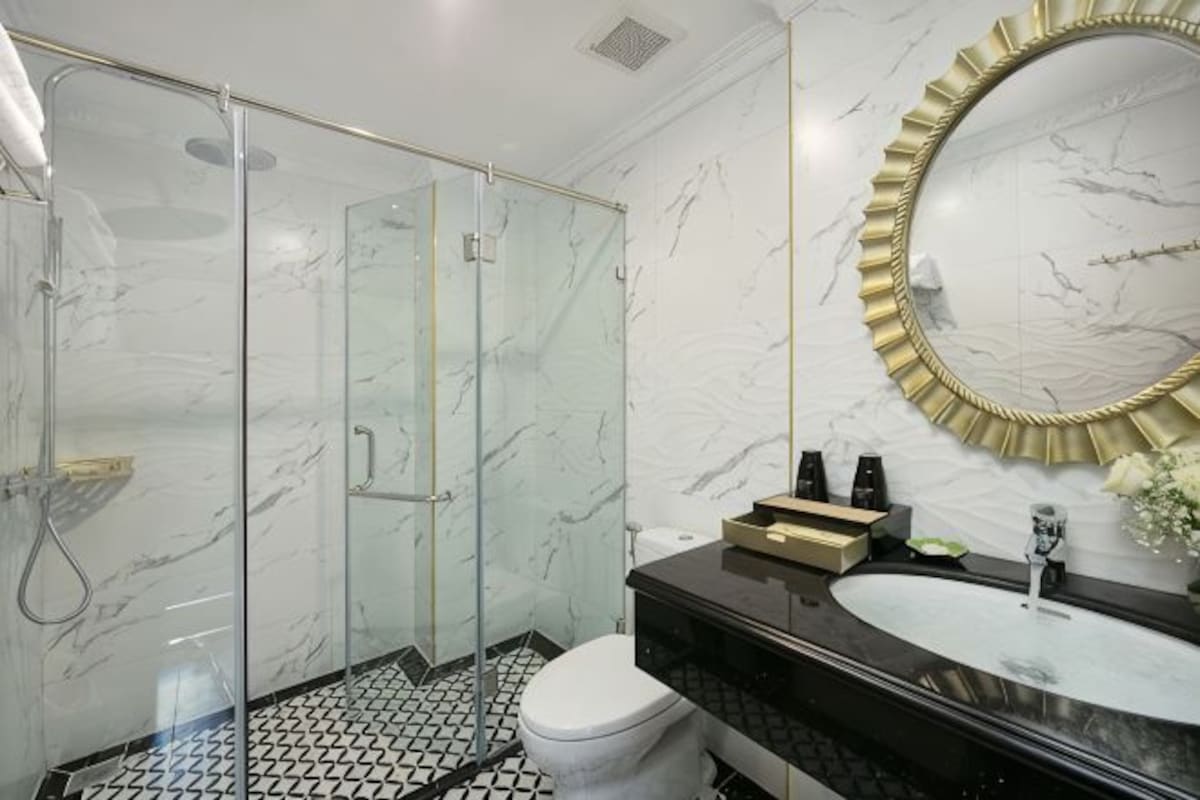 A bathroom with a glass shower enclosure, a marble wall, a round mirror with a gold frame, and a black countertop with a sink.