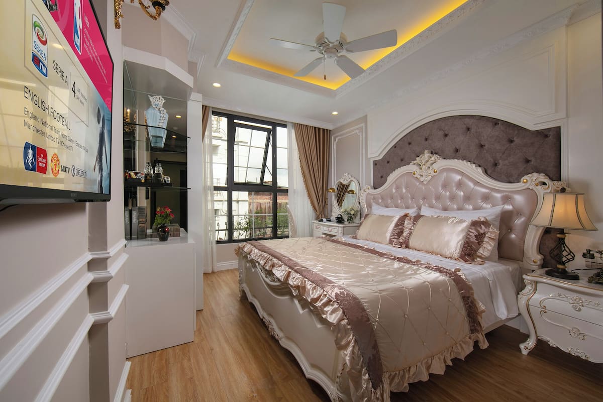 A bedroom with a large bed, a ceiling fan, a window with curtains, and a TV displaying sports logos.