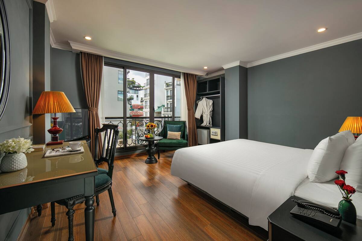 A hotel room with a bed, a desk with a lamp, a chair, a small table with a vase of flowers, and a view of buildings through the window.