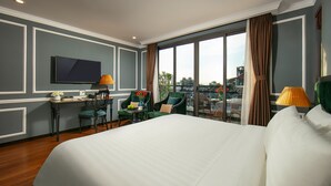 Ein Hotelzimmer mit einem großen Bett, einem Schreibtisch mit Lampe, zwei Stühlen und Blick auf die Stadt durch große Fenster.