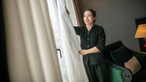 Eine Person steht an einem Fenster mit Vorhängen, ein grünes Sofa mit Kissen und eine Lampe auf einem Beistelltisch.