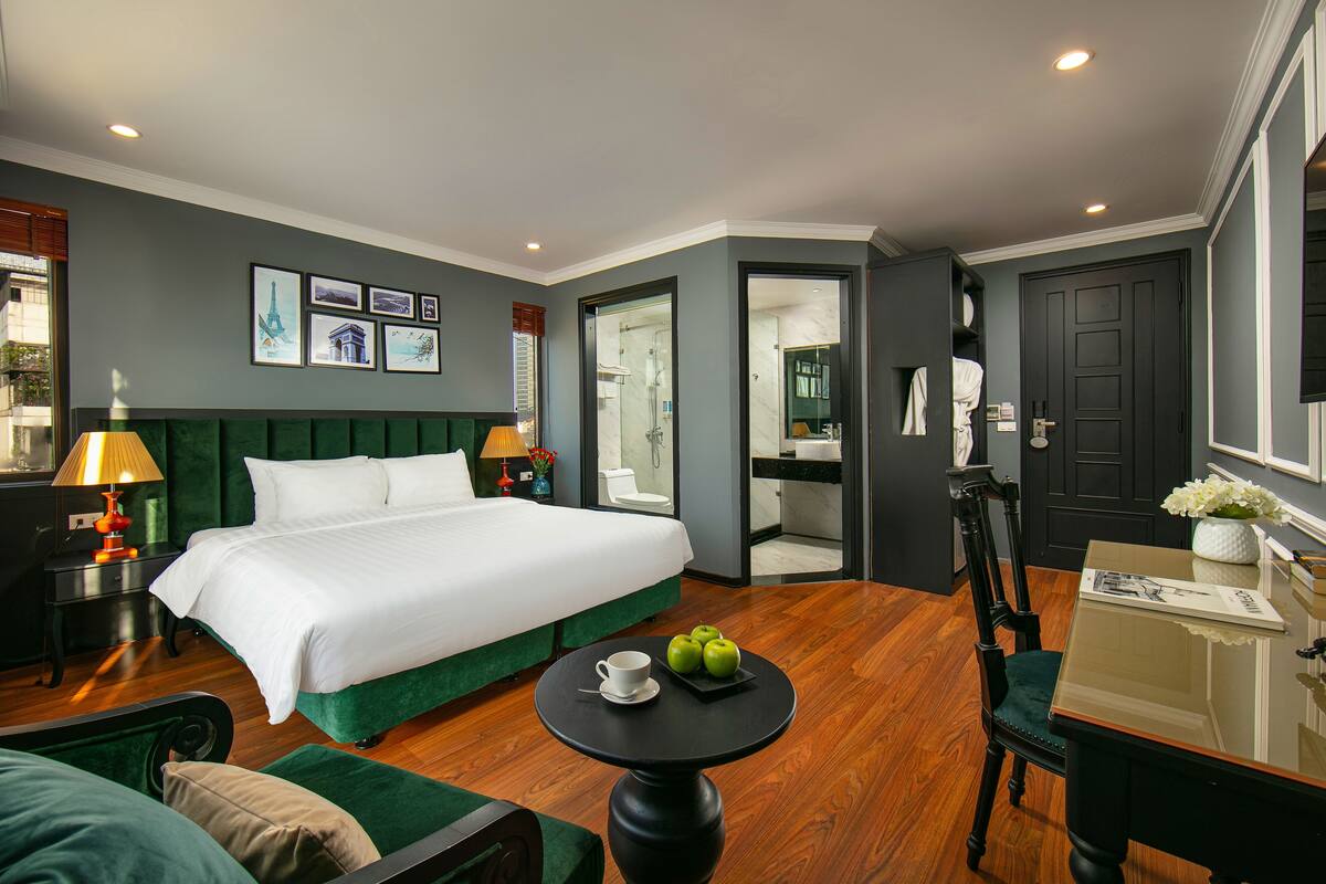 A hotel room with a large bed, a small round table with apples and a cup, a chair, a desk with a mirror, and a door leading to a bathroom.