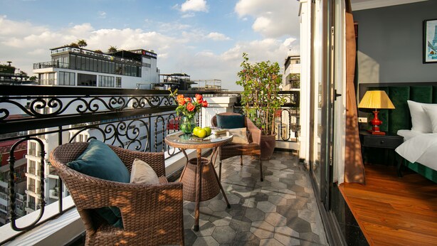Ein Balkon mit Korbmöbeln, einem Tisch mit Obst und Blumen sowie Blick auf die Stadtlandschaft.