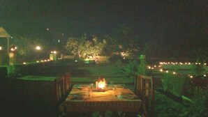 Un espacio de comedor al aire libre con una mesa y sillas, un farol iluminado y vistas a un edificio lejano y árboles.