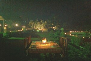 An outdoor dining area with a table and chairs, a lit lantern, and a view of a distant building and trees.