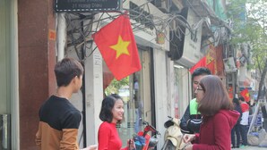 A street scene with people standing and interacting. Flags are visible, and a scooter is parked on the sidewalk. - Rendez Vous Hotel (Hanoi)