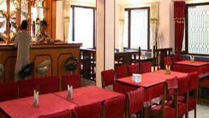 Ein Restaurant mit roten Tischdecken und Stühlen, einer Theke mit einer Speisekarte und großen Fenstern mit Vorhängen.